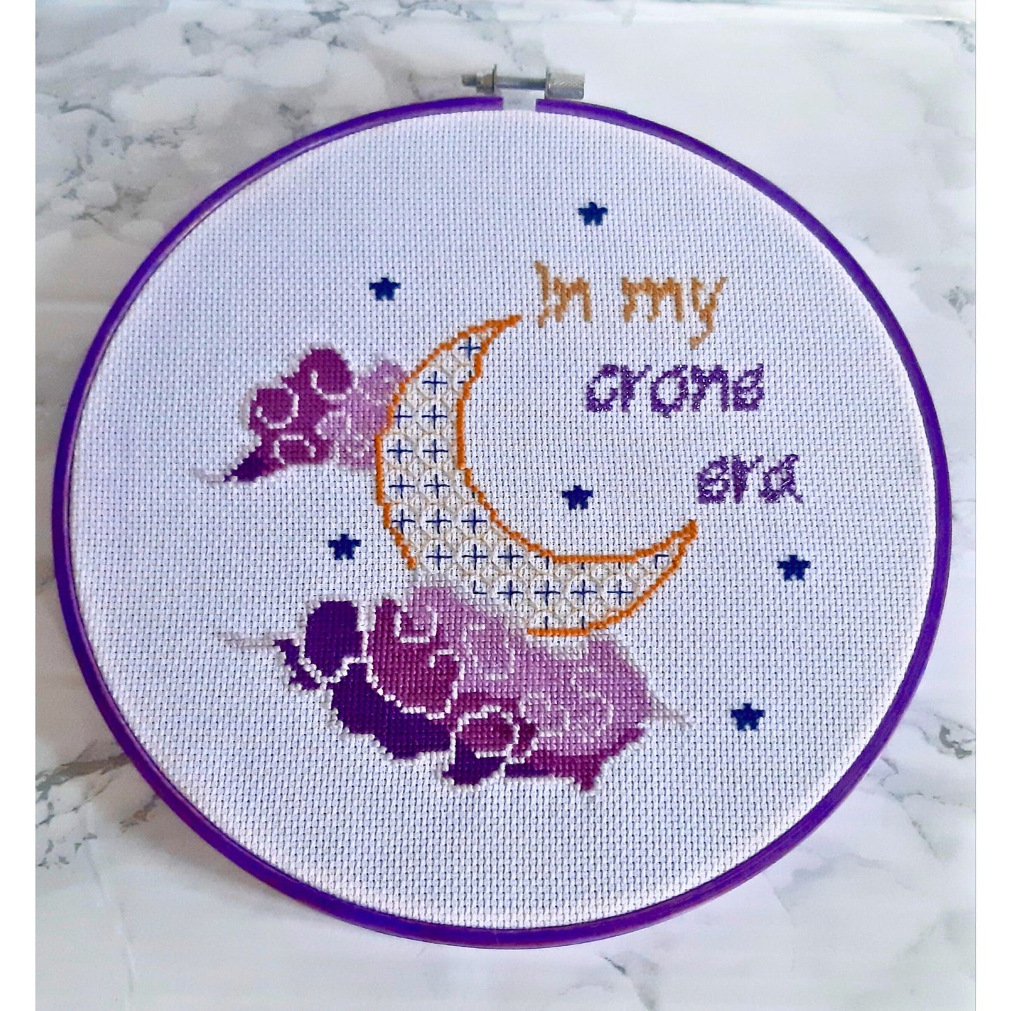 Completed cross stitch quote in an 8 inch hand dyed purple embroidery hoop. Features a moon and clouds motif and the words 'In my crone era' The piece is made up of purples and gold threadhoop