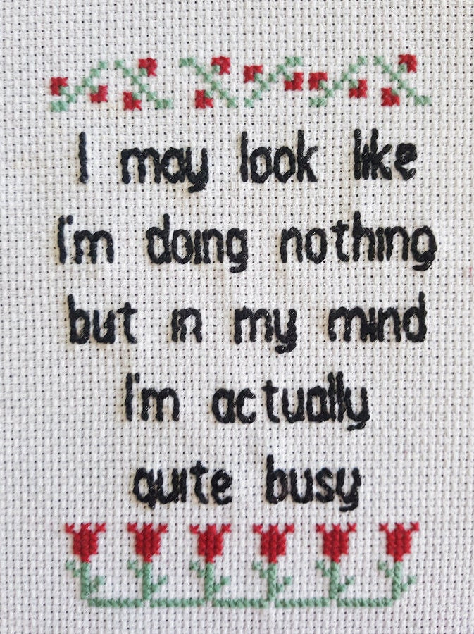 Close up of completed embroidery quote with black text on white background surrounded by red and green flowers. Text says 'I may look like I'm doing nothing but in my mind I'm actually quite busy'
