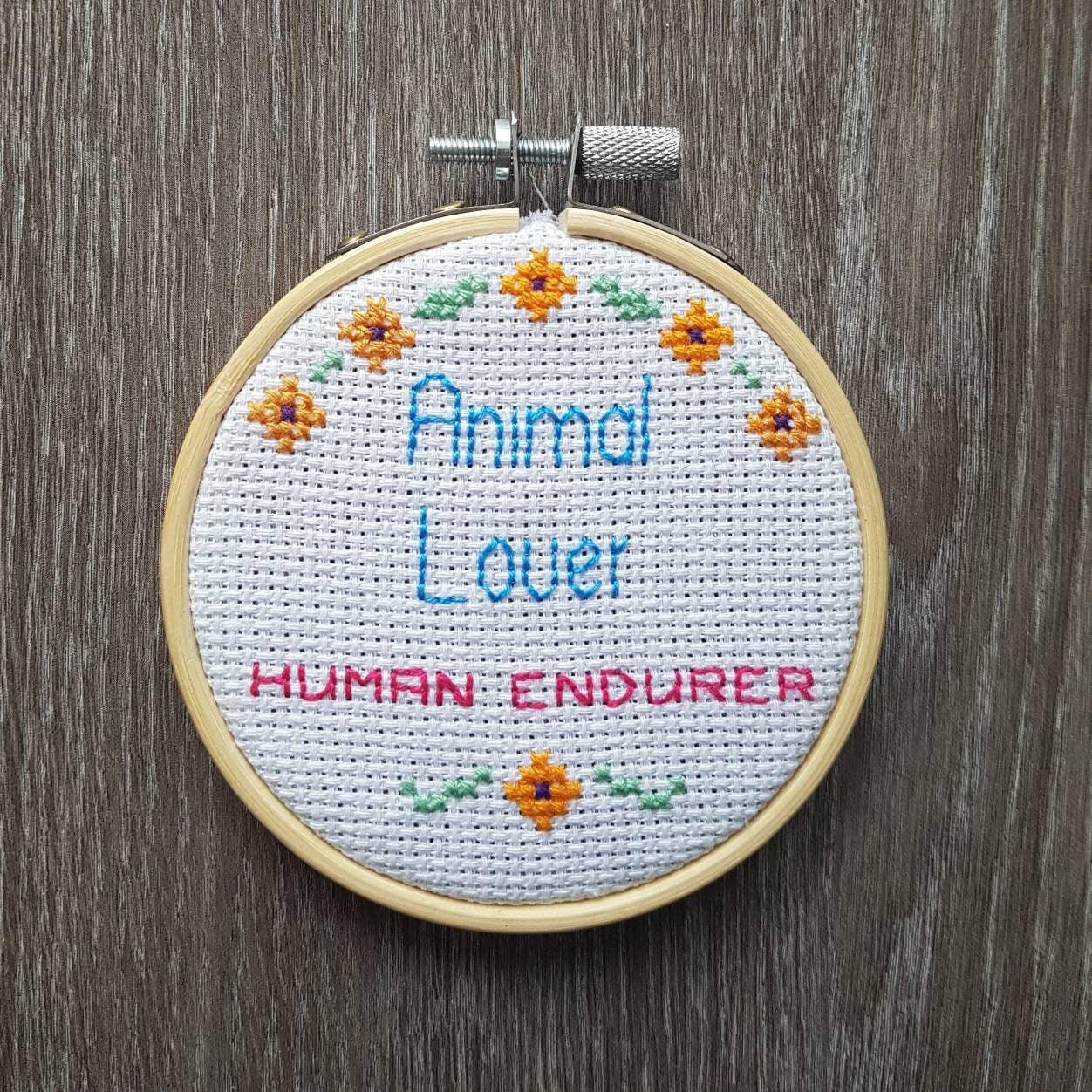 Animal lover, human endurer cross stitch quote in 3 inch embroidery hoop on dark wood background. Quote is surrounded by small flowers
