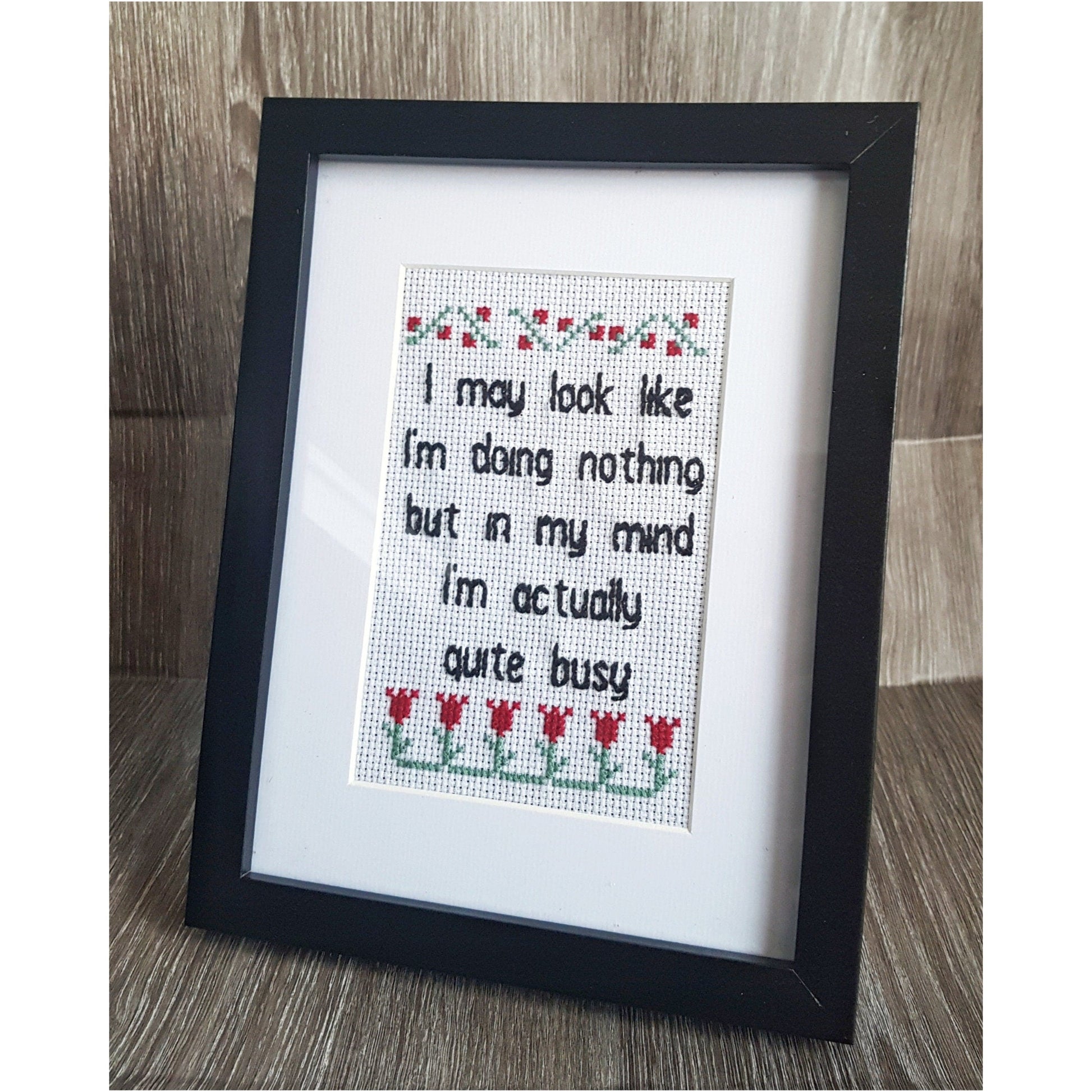 Completed embroidery quote with black text on white background surrounded by red and green flowers. Text says 'I may look like I'm doing nothing but in my mind I'm actually quite busy'. Displayed in black frame with white mount