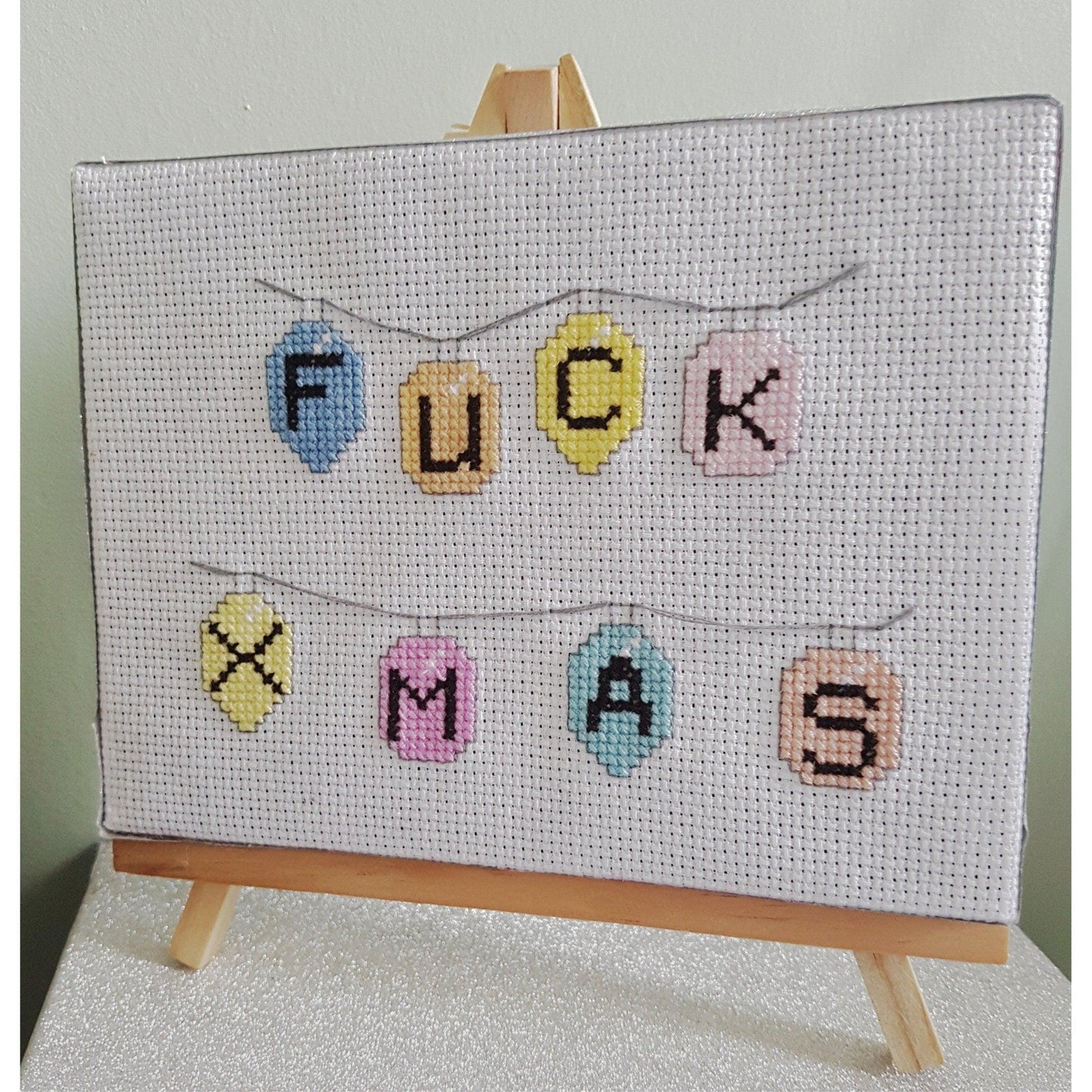 Completed cross stitch on canvas and easel with the words 'Fuck Xmas'