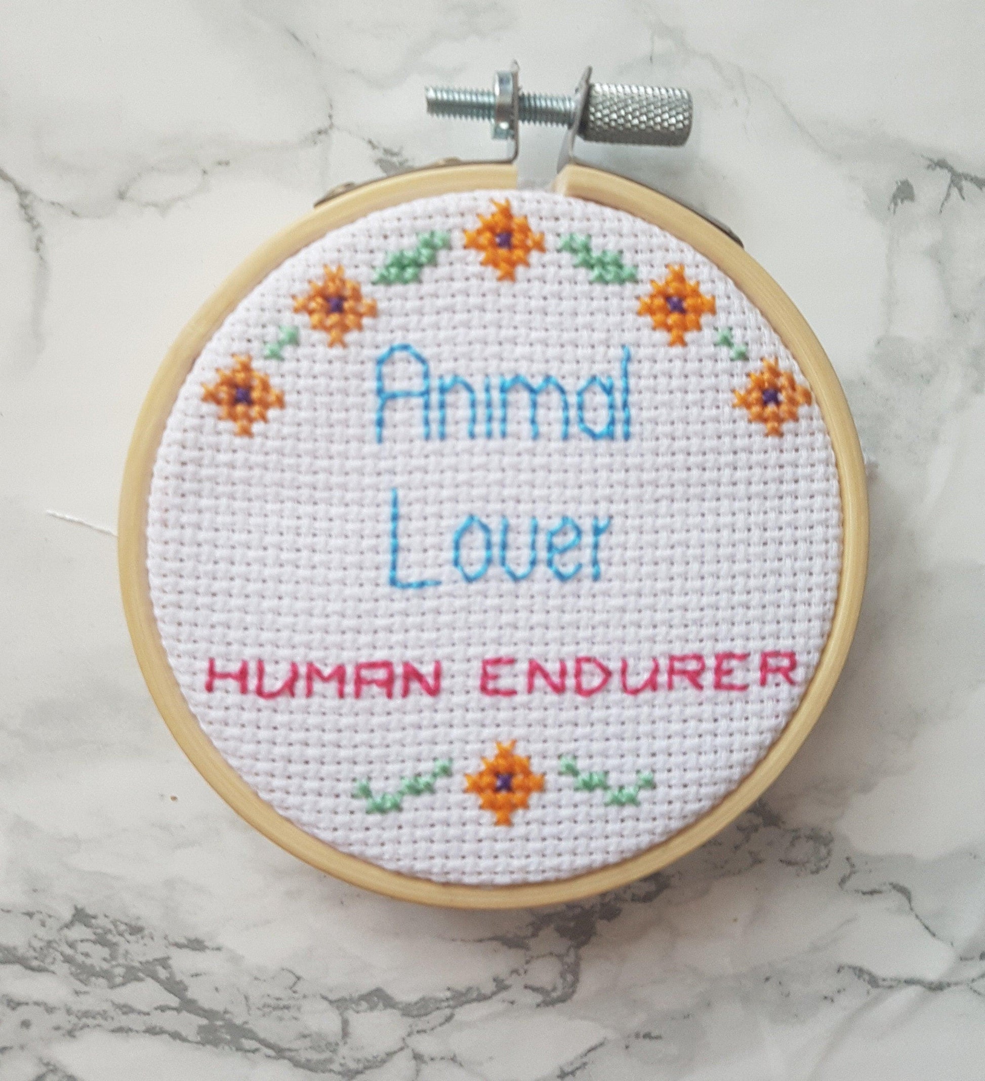 Animal lover, human endurer cross stitch quote in 3 inch embroidery hoop on white marbled background. Quote is surrounded by small flowers