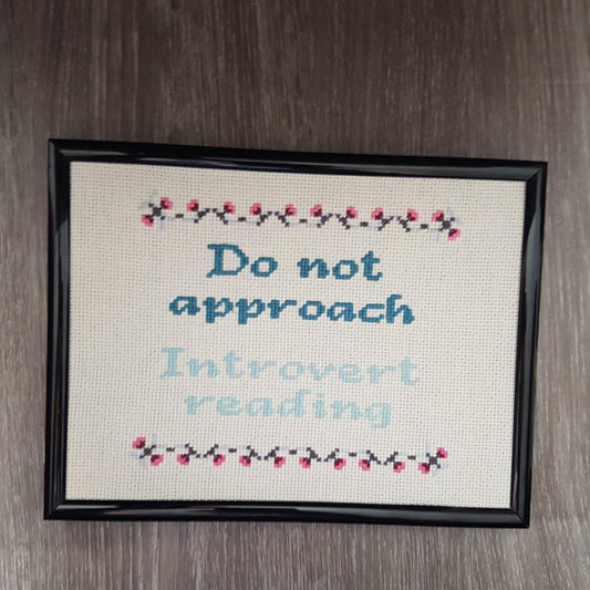 Completed cross stitch quote with the text 'Do not approach, introvert reading' in light and dark blue-green thread. Quote has a grey and pink border of small flowers. Cross stitch has a black frame   