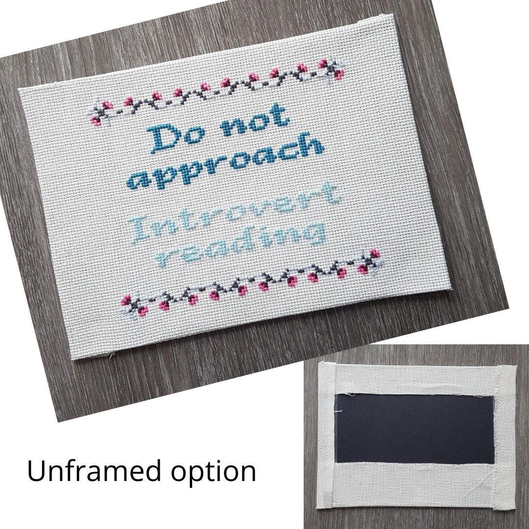 Completed cross stitch quote with the text 'Do not approach, introvert reading' in light and dark blue-green thread. Quote has a grey and pink border of small flowers. Cross stitch has no frame and is mounted on black card  