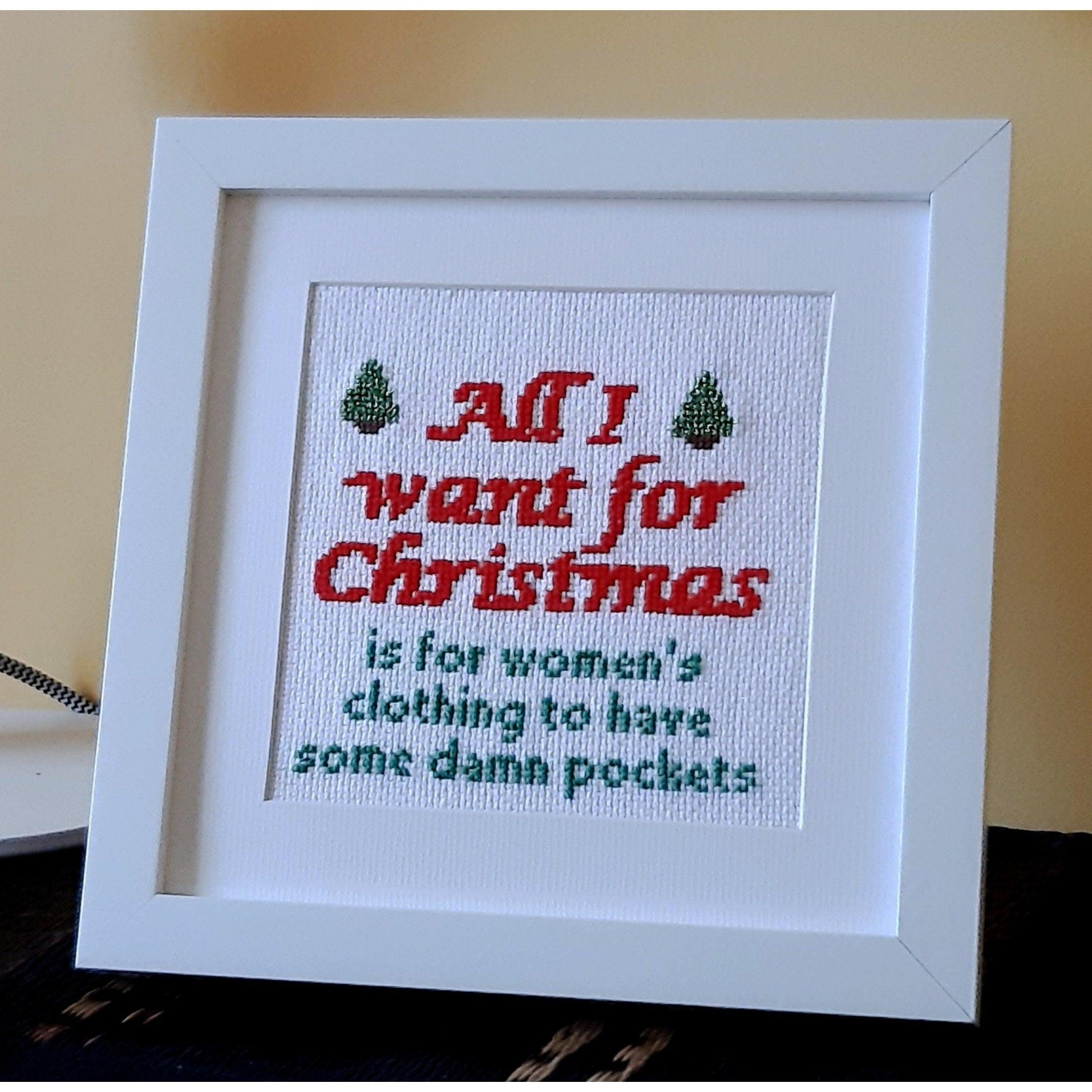 Completed Christmas cross stitch in chunky white frame saying 'All I want for Christmad is for women's clothing to have some damn pockets'