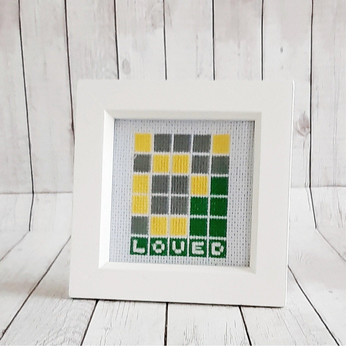 Completed cross stitch showing a wordle game with the solution being the word 'loved'. It is on white fabric, with a white square chunky frame
