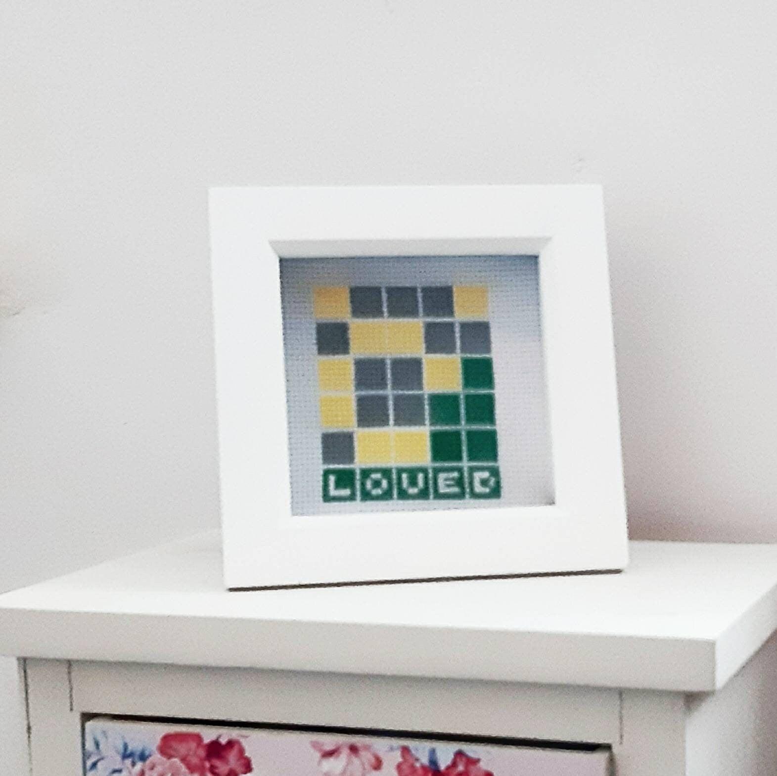 Completed cross stitch showing a wordle game with the solution being the word 'loved'. It is on white fabric, with a white square chunky frame. Frame is positioned on drawers to illustrate size